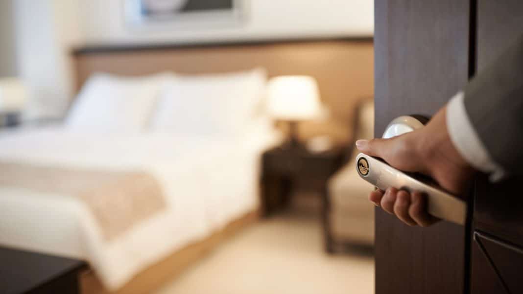 Widok na hotelowy pokój z zamkniętymi drzwiami, w tle łóżko i lampka nocna.