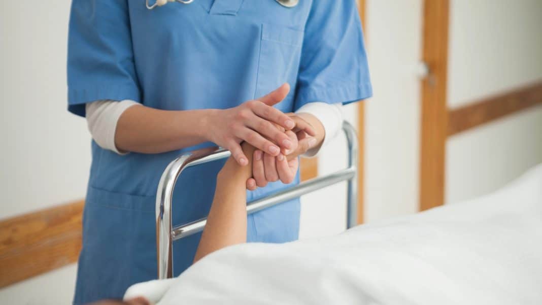 Zdjęcie pielęgniarki trzymającej rękę pacjenta w szpitalu, widoczna część łóżka i personelu medycznego.