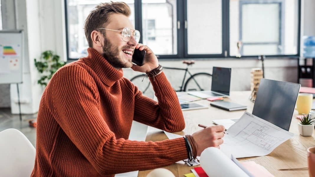 Mężczyzna rozmawia przez telefon w biurze, siedząc przy stole z laptopem i dokumentami.