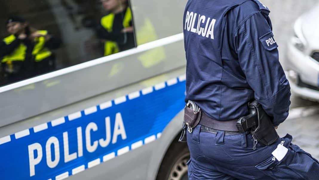 Policjant w uniformie obok radiowozu na ulicy, widoczne odbicie w lusterku samochodu.