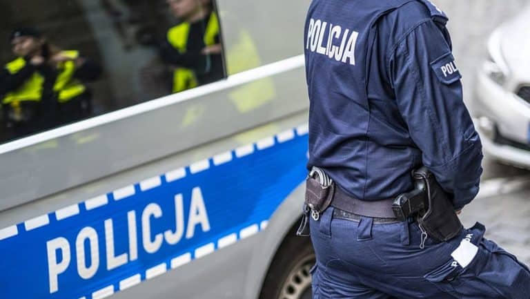 Policjant w uniformie obok radiowozu na ulicy, widoczne odbicie w lusterku samochodu.