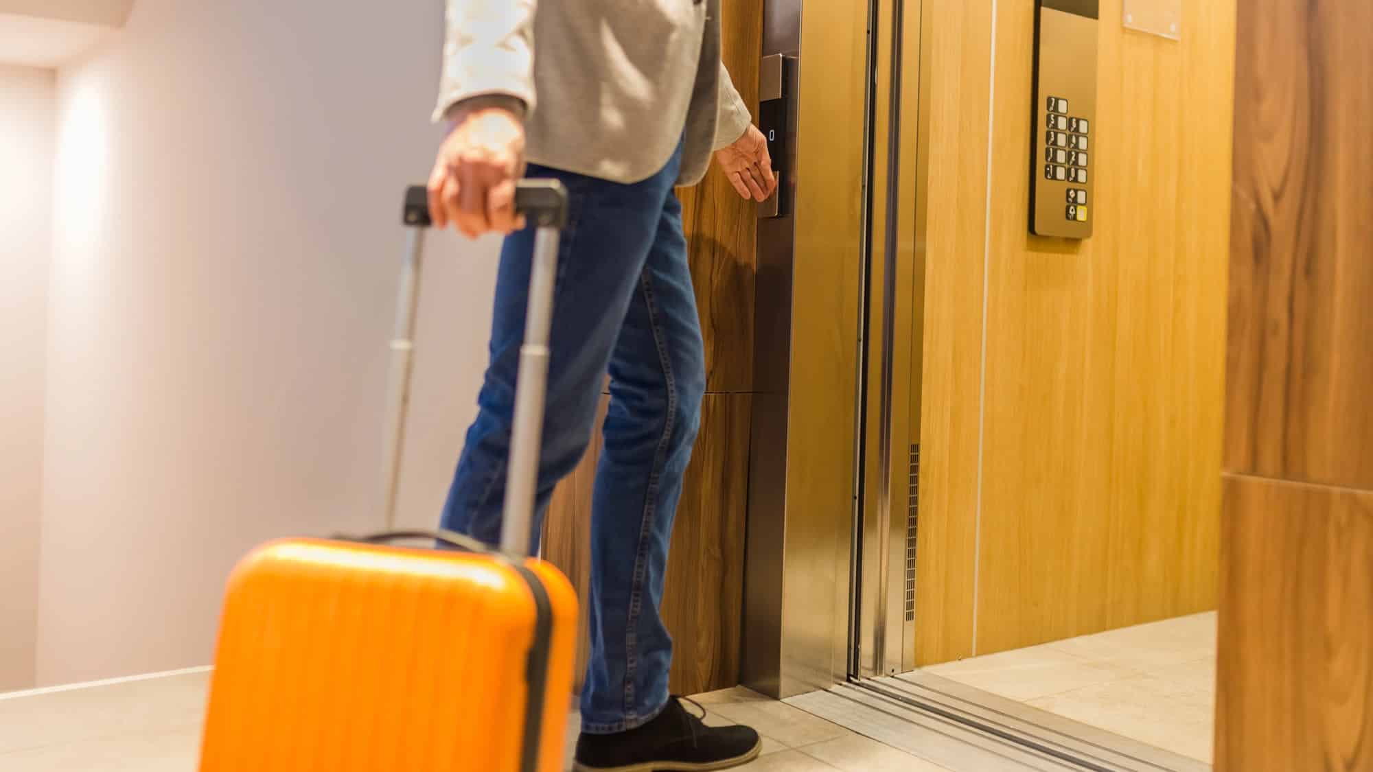Osoba z walizką wjeżdża windą w budynku hotelowym lub apartamentowym.