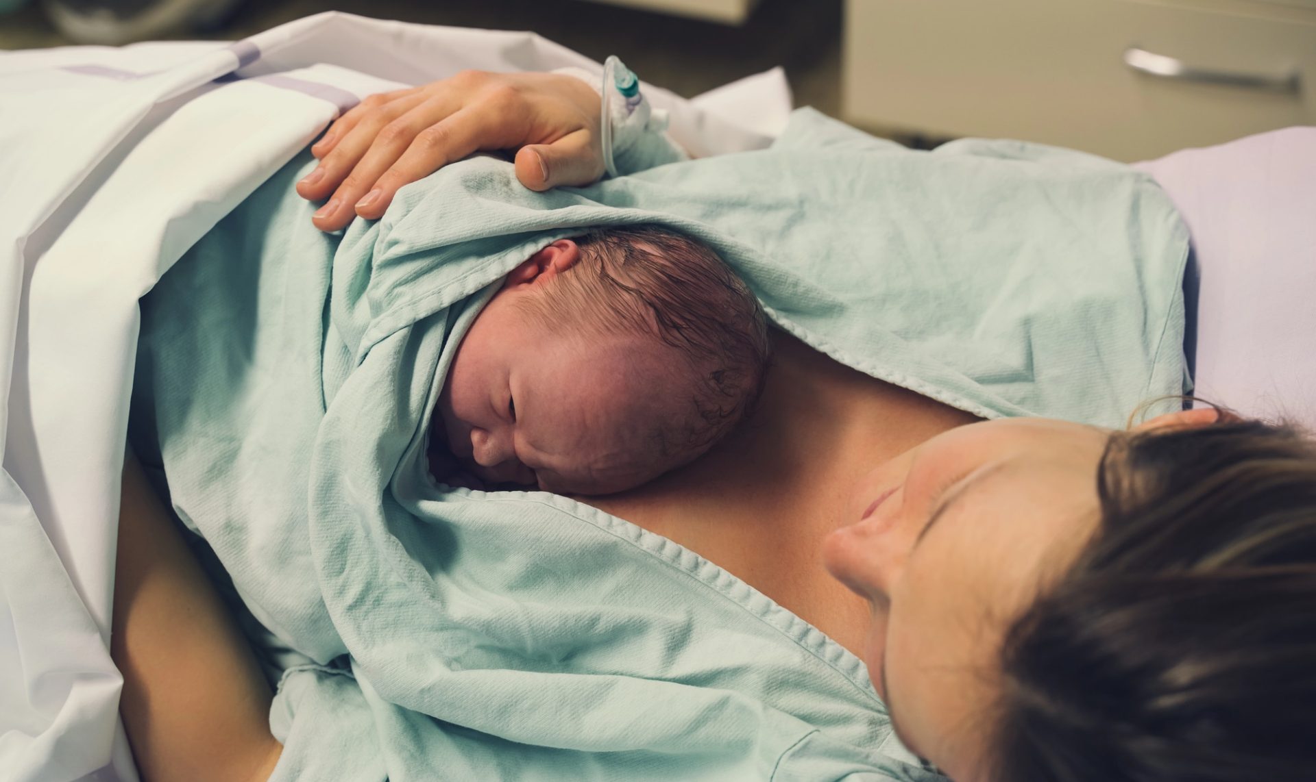 Kobieta trzyma noworodka na piersi, leżąc na łóżku w szpitalu.