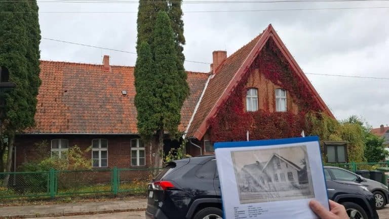 Osoba trzyma w ręku fotografię starego domu na tle obecnego budynku, na ulicy z samochodami.