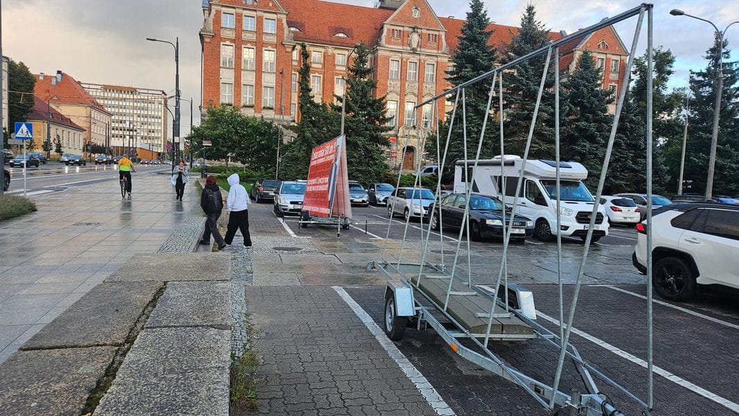 Baner z wizerunkiem wiceprezydent Olsztyna zabezpieczony przez policję, ustawiony na chodniku obok parkingu.