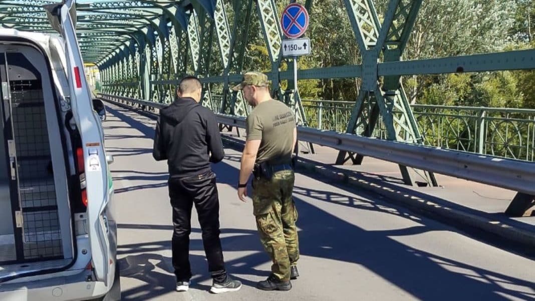 Dwóch mężczyzn rozmawia na moście granicznym, jeden w mundurze, drugi w casualowym ubraniu.