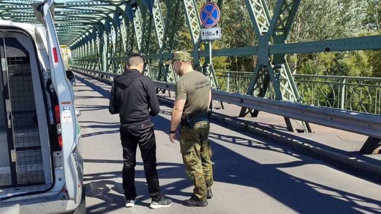 Dwóch mężczyzn rozmawia na moście granicznym, jeden w mundurze, drugi w casualowym ubraniu.