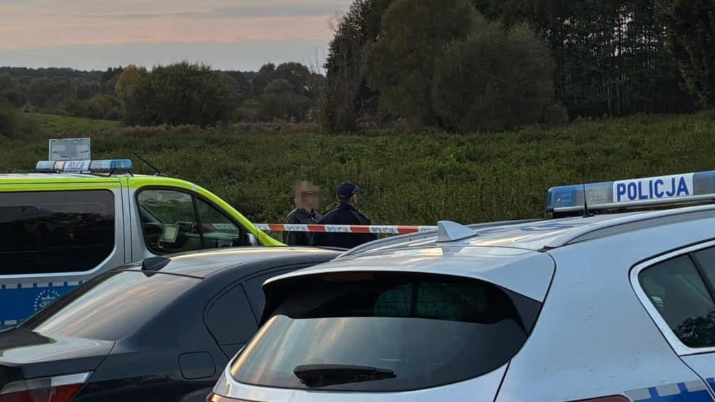 Policjanci rozmawiają na łące, otoczeni samochodami policyjnymi, w trakcie akcji poszukiwawczej.