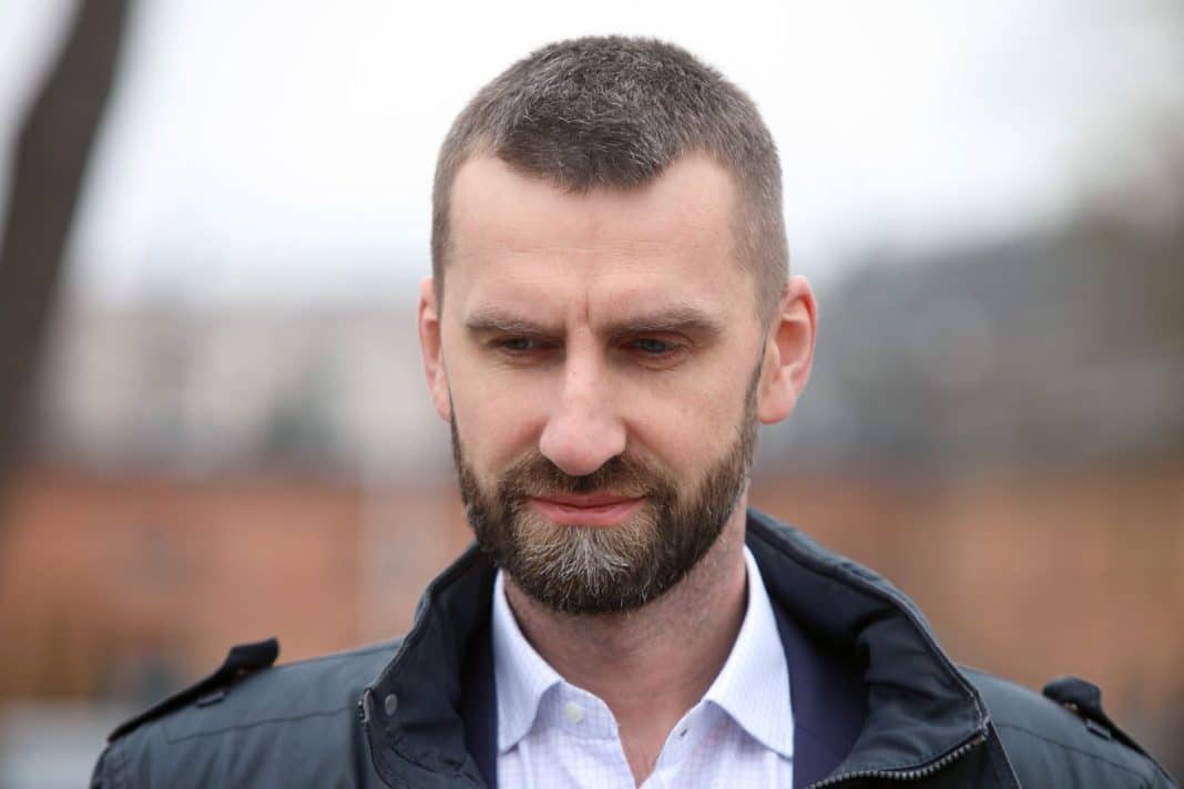 Zdjęcie mężczyzny w ciemnej kurtce i białej koszuli na tle nieba i budynków.