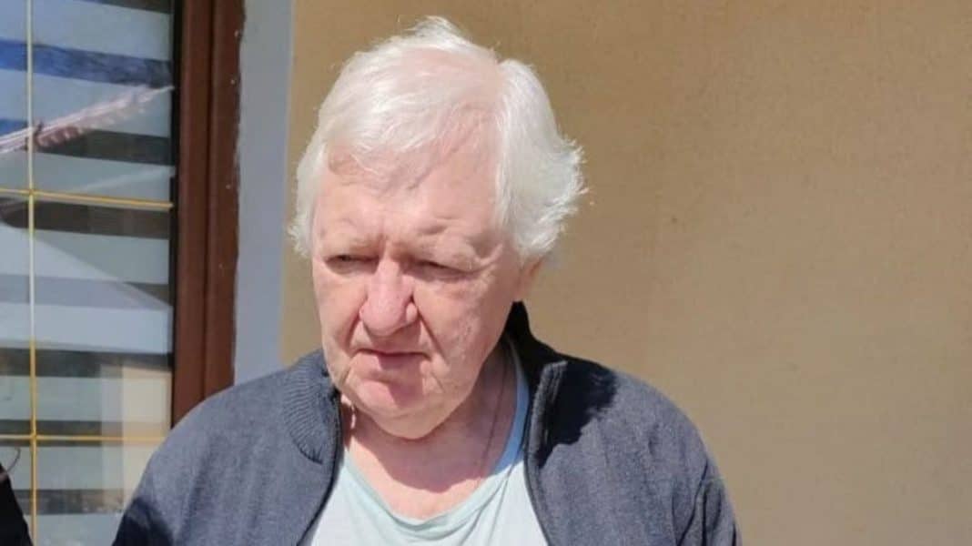 Zdjęcie starszego mężczyzny z białymi włosami, ubrany w jasną koszulkę i ciemną kurtkę, stojącego na zewnątrz.