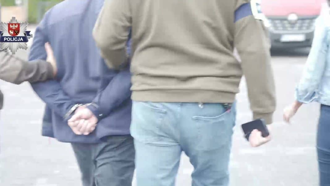 Grupa mężczyzn zatrzymanych przez policję na ulicy, widoczne ich plecy, w tle samochody.