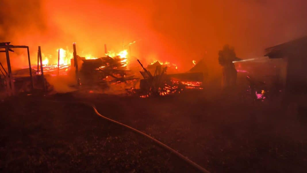 Pożar kompleksu budynków gospodarczych w nocy, widoczne płomienie i dym.
