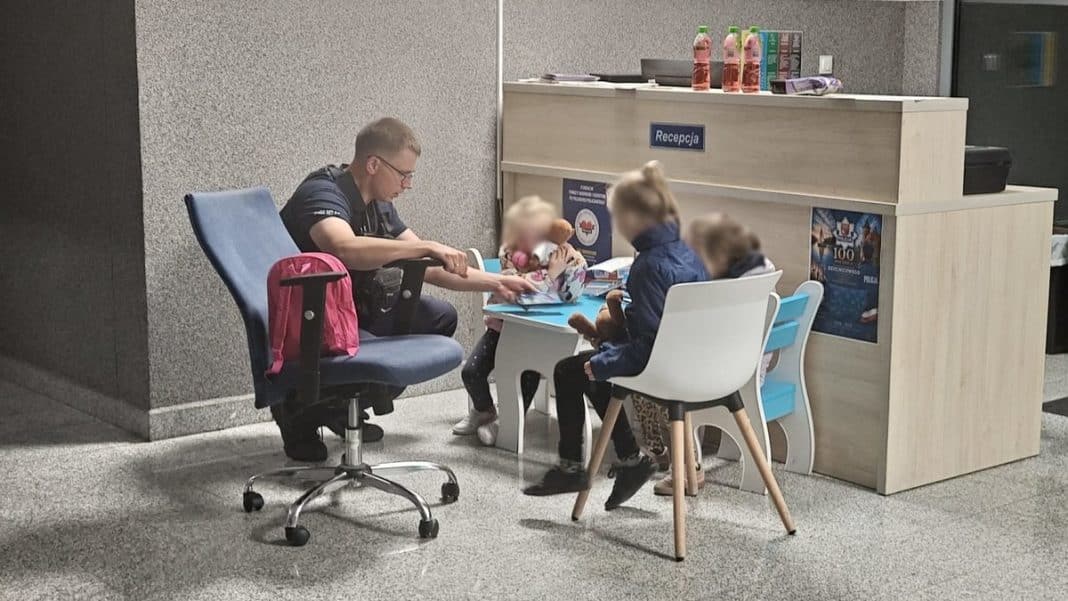Policjant rozmawia z trzema dziećmi w poczekalni, obok recepcja, w tle szara ściana.