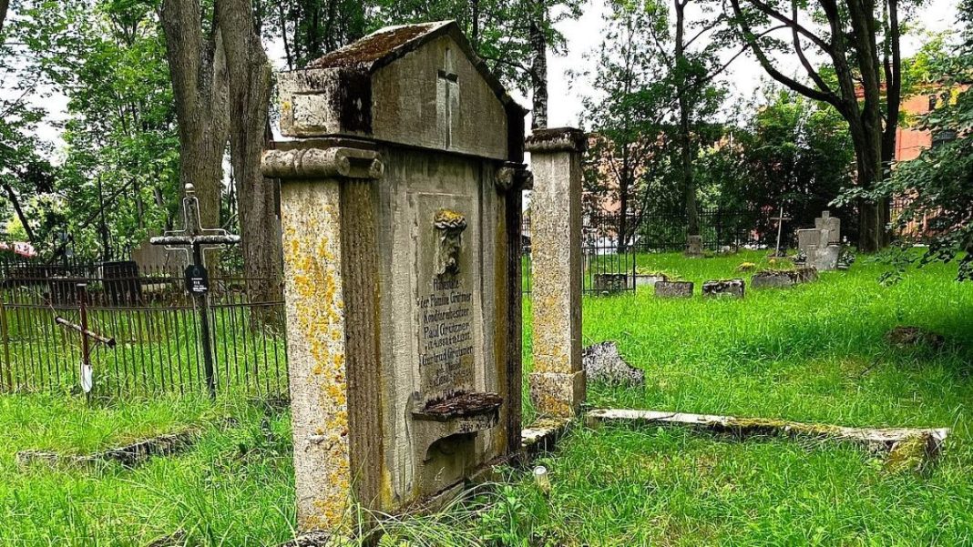 Stary nagrobek na cmentarzu św. Jakuba w Olsztynie, otoczony zielenią i innymi zabytkami nagrobków.