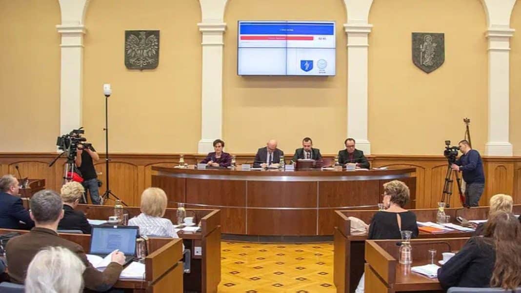 Sala sesyjna z czterema osobami na mównicy i kilkoma radnymi, na ścianie wyświetlacz z wynikami głosowania.