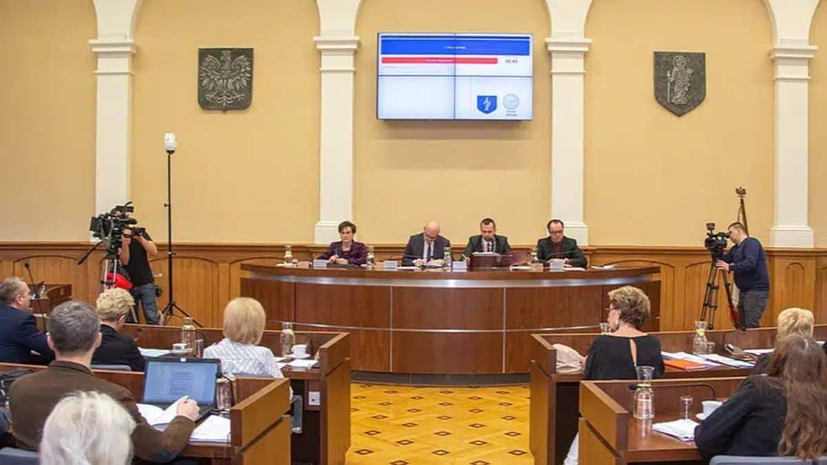 Sala sesyjna z czterema osobami na mównicy i kilkoma radnymi, na ścianie wyświetlacz z wynikami głosowania.
