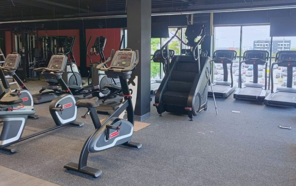 Wnętrze nowoczesnej siłowni z bieżniami, rowerami i innym sprzętem fitness, duże okna z widokiem na miasto.