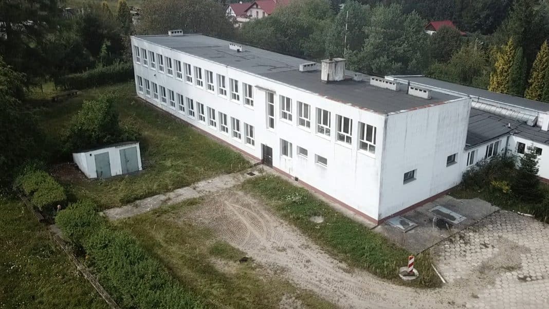 Widok na nieczynną szkołę z ukrytym pod nią schronem z czasów PRL, otoczony zielenią i domami.