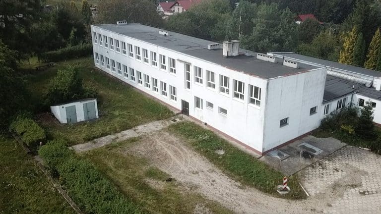 Widok na nieczynną szkołę z ukrytym pod nią schronem z czasów PRL, otoczony zielenią i domami.
