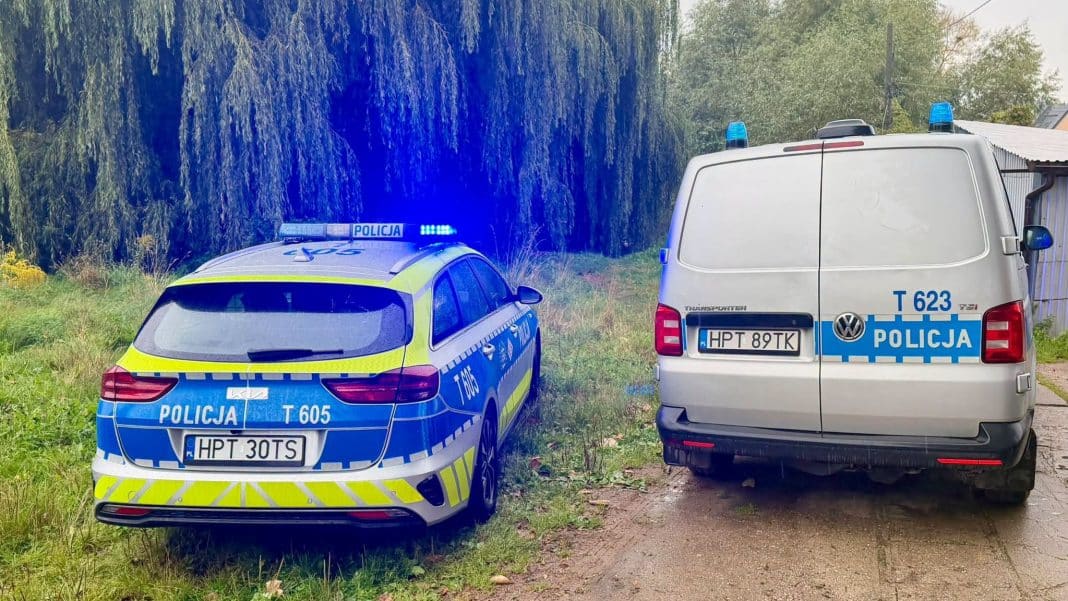Dwa radiowozy policyjne stoją na poboczu przy zielonym terenie, jeden z nich z włączonymi światłami.