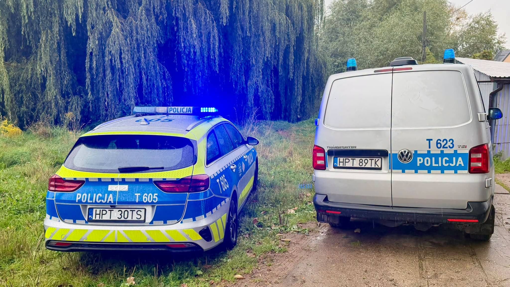 Dwa radiowozy policyjne stoją na poboczu przy zielonym terenie, jeden z nich z włączonymi światłami.