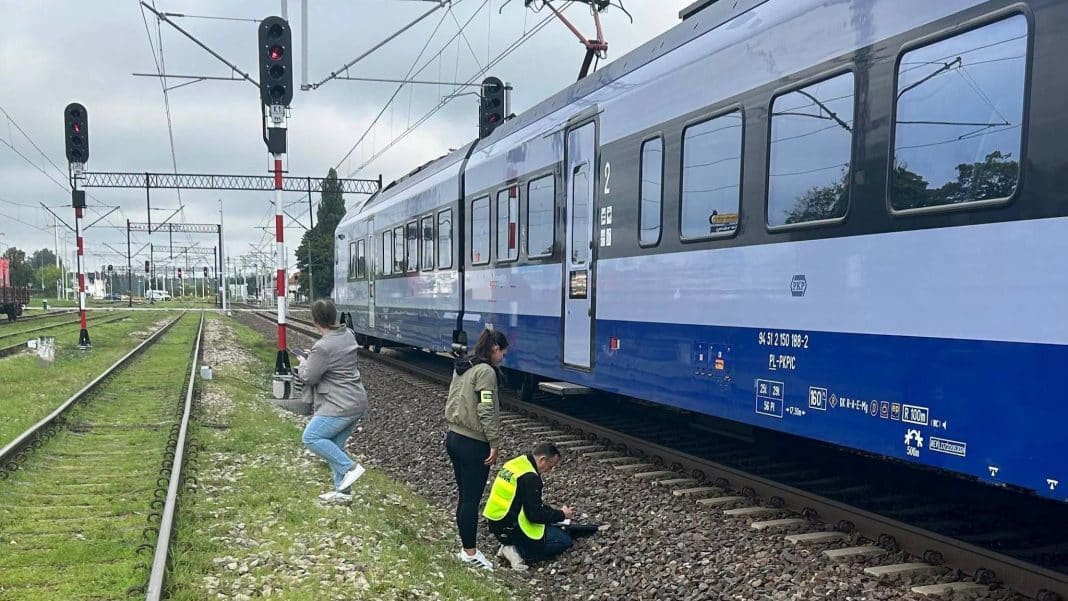 Na zdjęciu pociąg stojący na torach w pobliżu stacji kolejowej, z grupą osób w pobliżu, jedna osoba na kolanach.