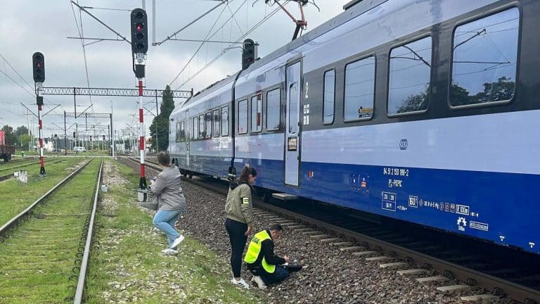 Na zdjęciu pociąg stojący na torach w pobliżu stacji kolejowej, z grupą osób w pobliżu, jedna osoba na kolanach.