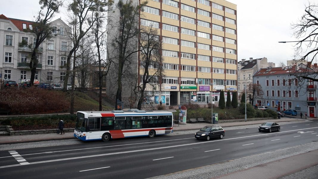 Autobus o nietypowym wyglądzie, pomalowany w biało-czerwone barwy, jedzie ulicą w mieście z wysokimi budynkami.