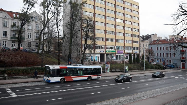 Autobus o nietypowym wyglądzie, pomalowany w biało-czerwone barwy, jedzie ulicą w mieście z wysokimi budynkami.