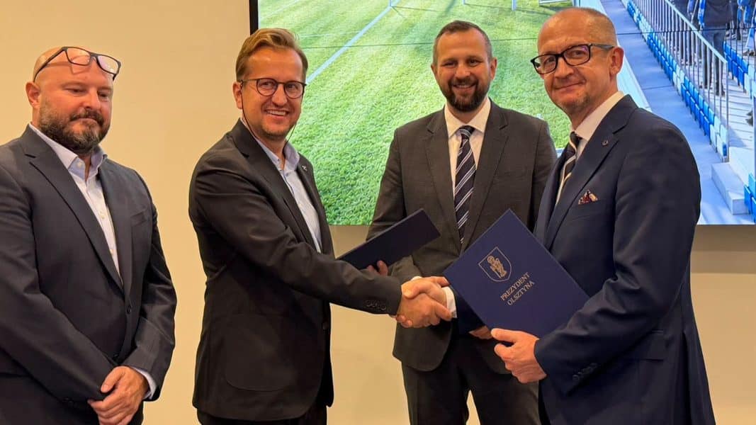 Czterech mężczyzn w garniturach podczas podpisania umowy, na ekranie w tle widać fragment stadionu.