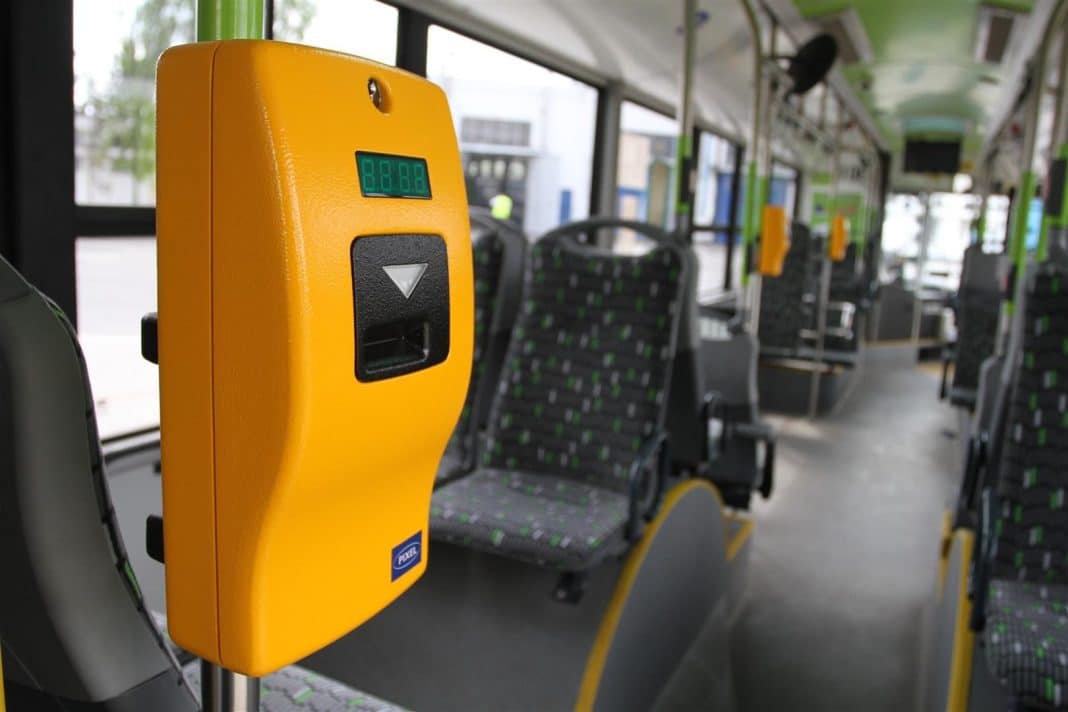 Żółty czytnik kart na tle wnętrza autobusu, w którym widać siedzenia i okna.