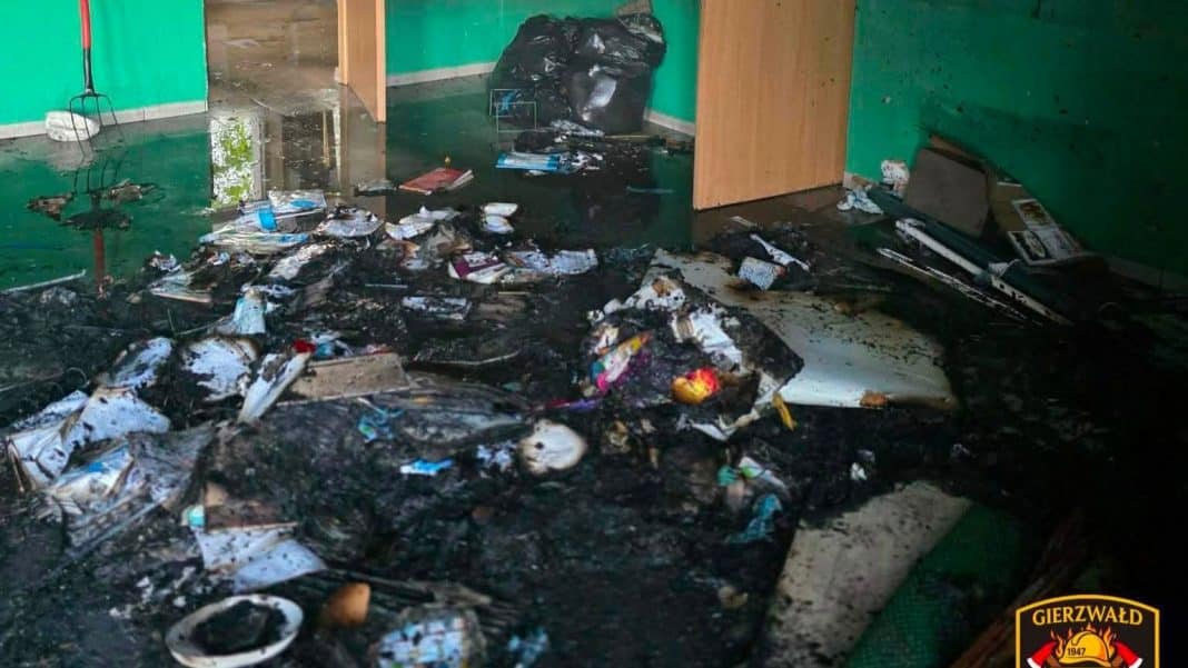 Zdewastowane wnętrze szkoły z pożarem i zniszczonymi meblami, oknami i instalacjami.
