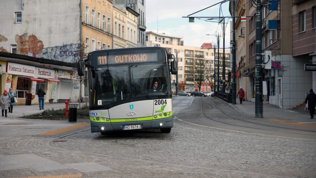 Autobus miejski na ulicy w mieście, w tle budynki i tramwaje, wczesny wieczór.