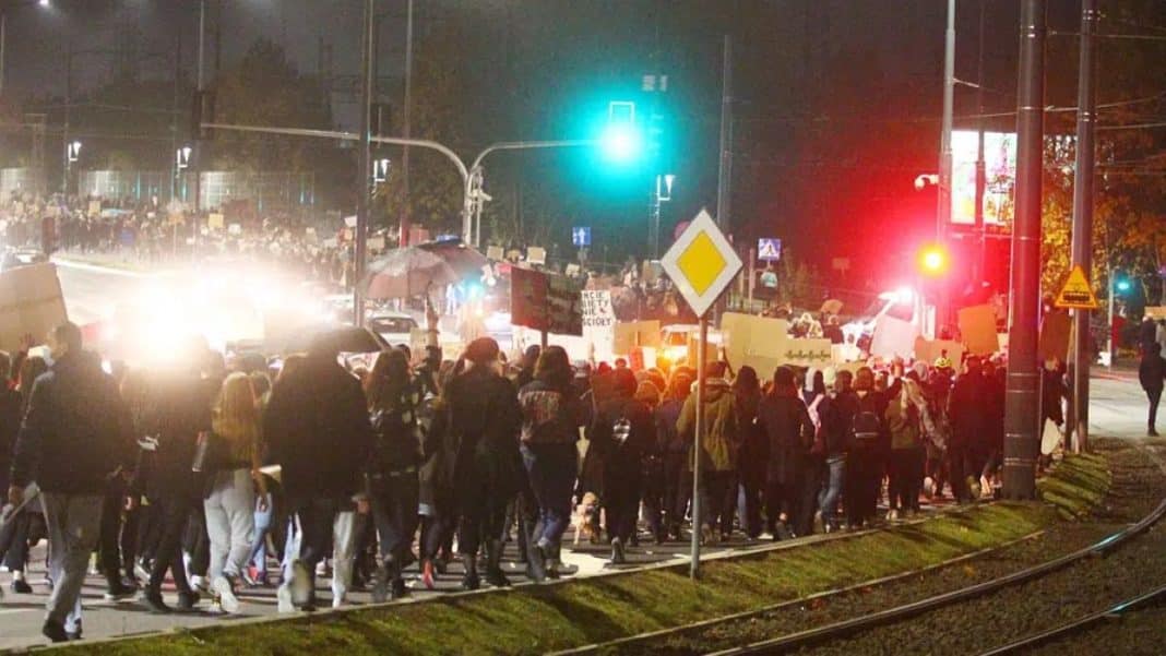 Grupa ludzi na ulicy podczas protestu z czerwonymi kartkami, wieczorem, w tle światła i tramwaje.