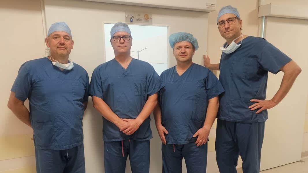 Grupa lekarzy w szpitalu, ubrani w niebieskie uniformy i czepki, stoją przed drzwiami oddziału.