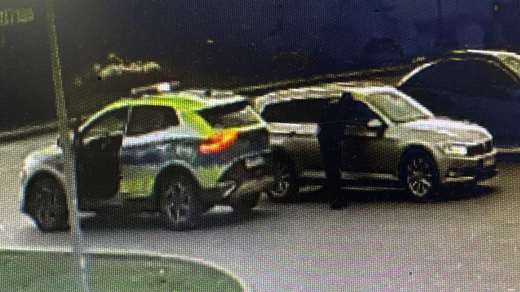 Zdjęcie przedstawia policyjny samochód i inny pojazd na parkingu, z funkcjonariuszem stojącym obok.
