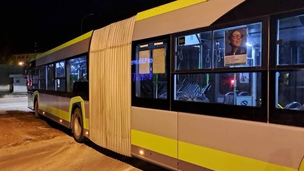 Autobus miejski na przystanku nocą, widoczny z boku, oświetlony wewnątrz i na zewnątrz, na tle ciemnego nieba.