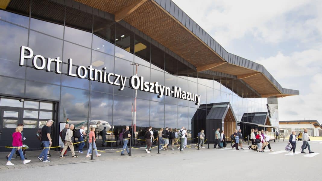 Grupa pasażerów wchodzących do terminalu lotniska z widocznym samolotem w tle.