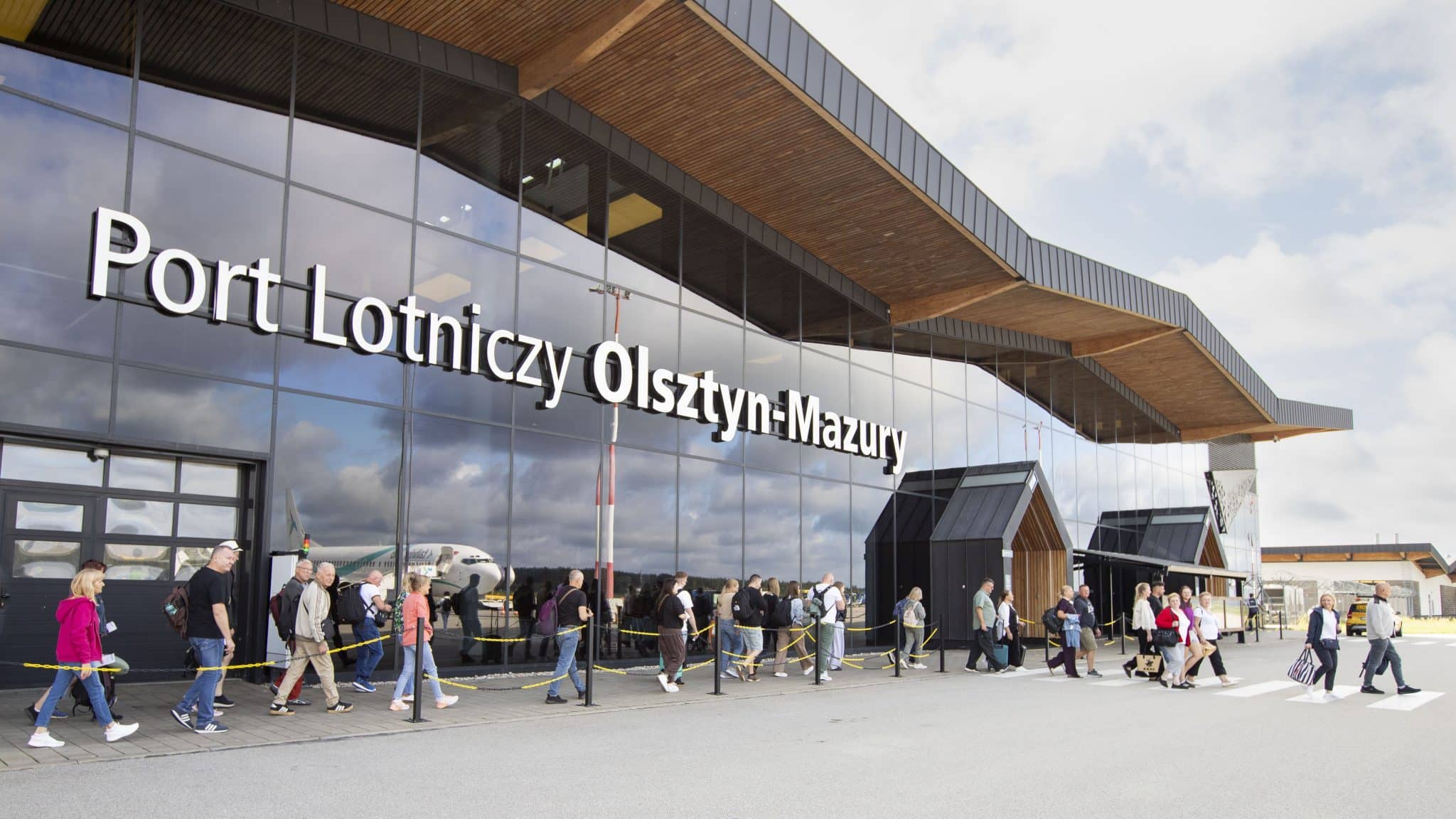 Grupa pasażerów wchodzących do terminalu lotniska z widocznym samolotem w tle.