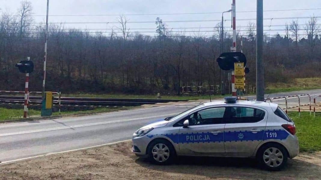 Samochód policyjny na przejeździe kolejowym z opuszczonymi rogatkami i sygnalizacją świetlną.