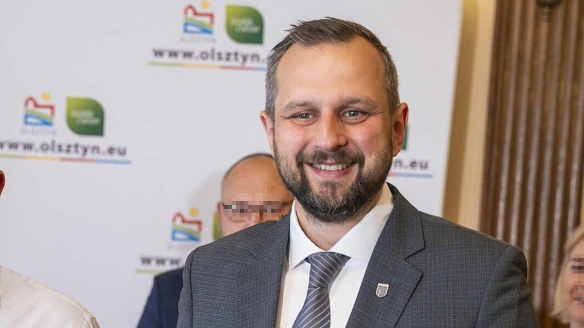 Mężczyzna w garniturze uśmiecha się na tle logo Olsztyna i innych osób.