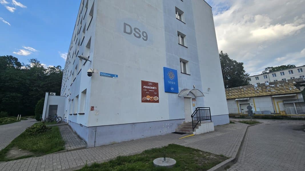Budynek wielopiętrowy z napisem D9 na ścianie i tablicami informacyjnymi, na tle chmur i drzew.