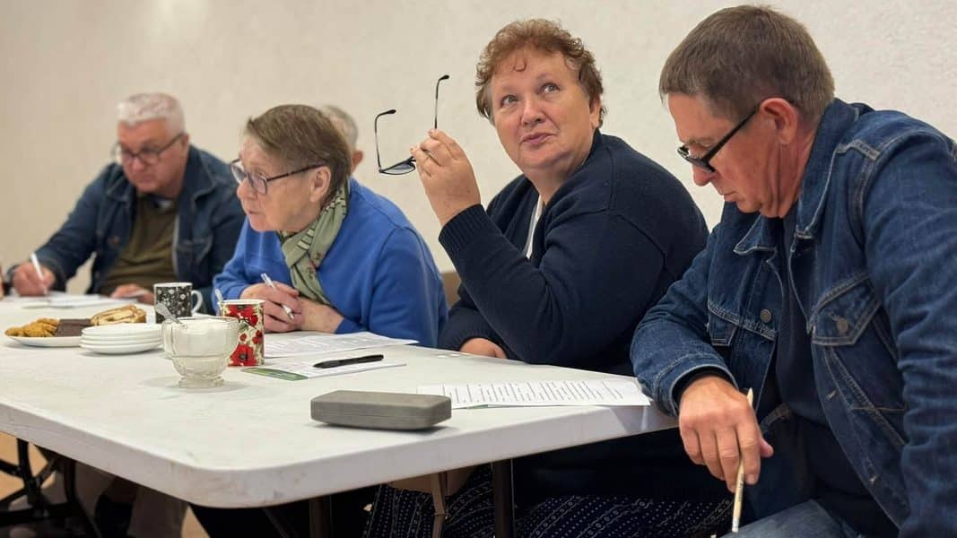 Grupa osób siedzi przy stole podczas spotkania, z kawą i ciastami na stole.