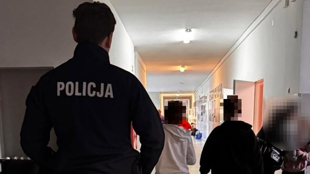 Policjant w szkole rozmawia z uczniami w korytarzu, w tle widoczne plakaty i inne dzieci.