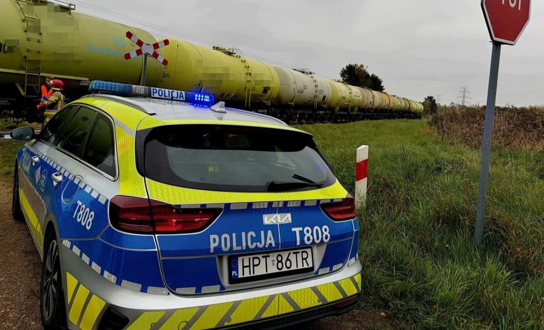Policja na miejscu wypadku przy torach kolejowych, pociąg i służby ratunkowe, w tle roślinność i niebo.