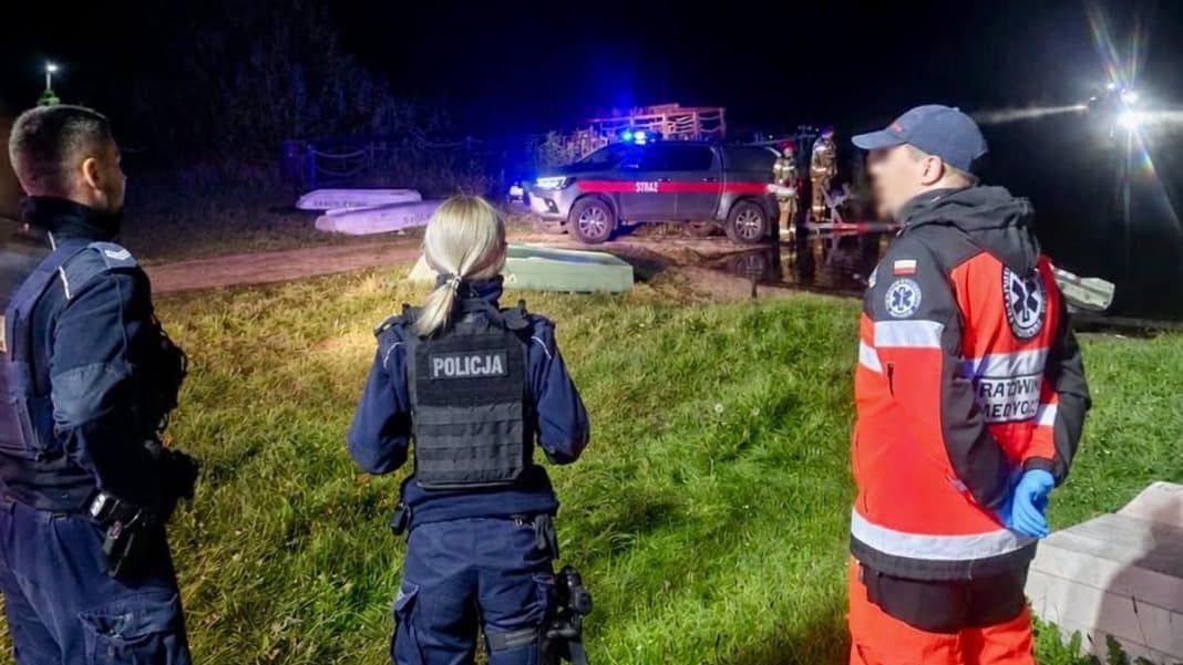 Służby ratunkowe i policja nad jeziorem podczas akcji nocnej, w tle samochód z sygnałami świetlnymi.
