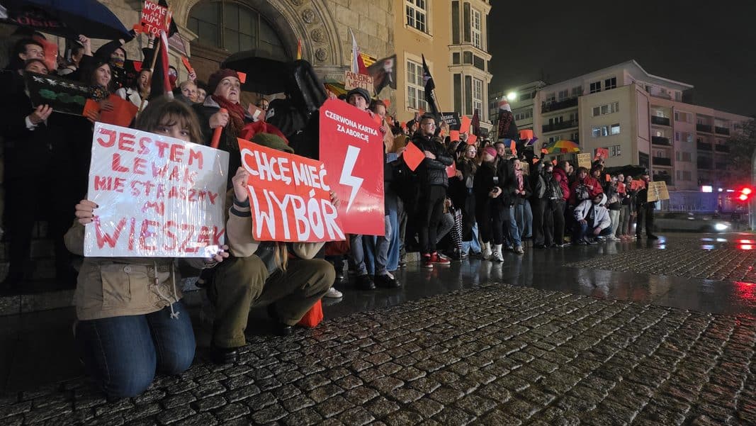 Grupa ludzi trzymających czerwone kartki i transparenty podczas protestu przed ratuszem w Olsztynie, wieczorem.