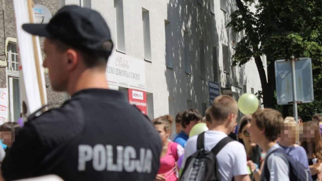Grupa uczniów stojących na zewnątrz szkoły, w tle widoczny policjant i budynek szkolny.