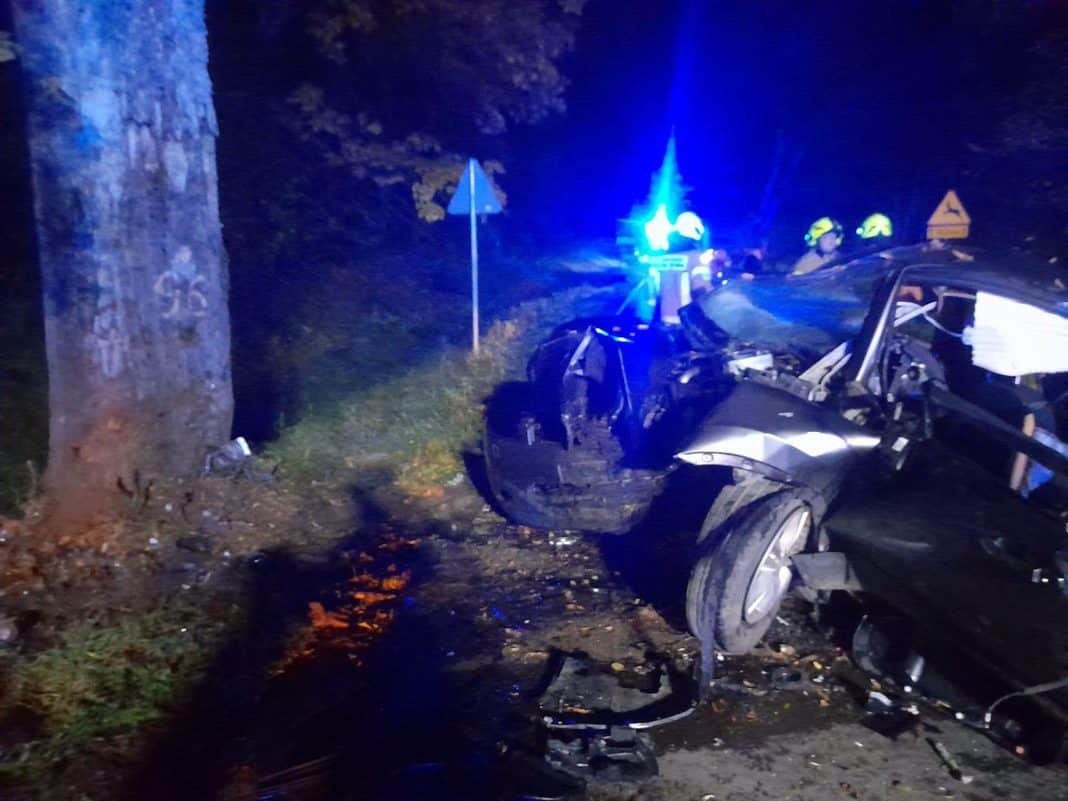 Rozbity samochód po wypadku na drodze wieczorem, w tle służby ratunkowe z sygnałami świetlnymi.