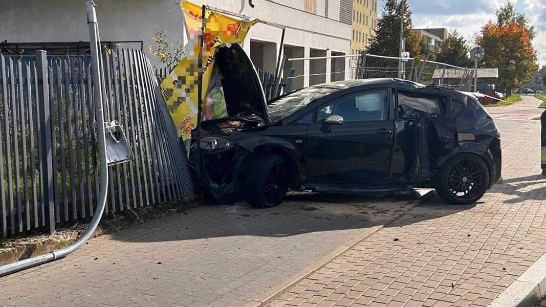 Uszkodzony czarny samochód po wypadku na chodniku, z uszkodzeniami z przodu i z boku, na tle ogrodzenia i budynków.
